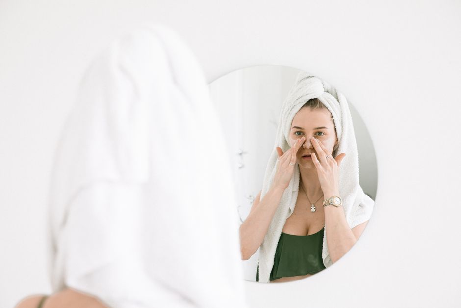 A woman in a towel performing her skincare routine in front of a bathroom mirror.